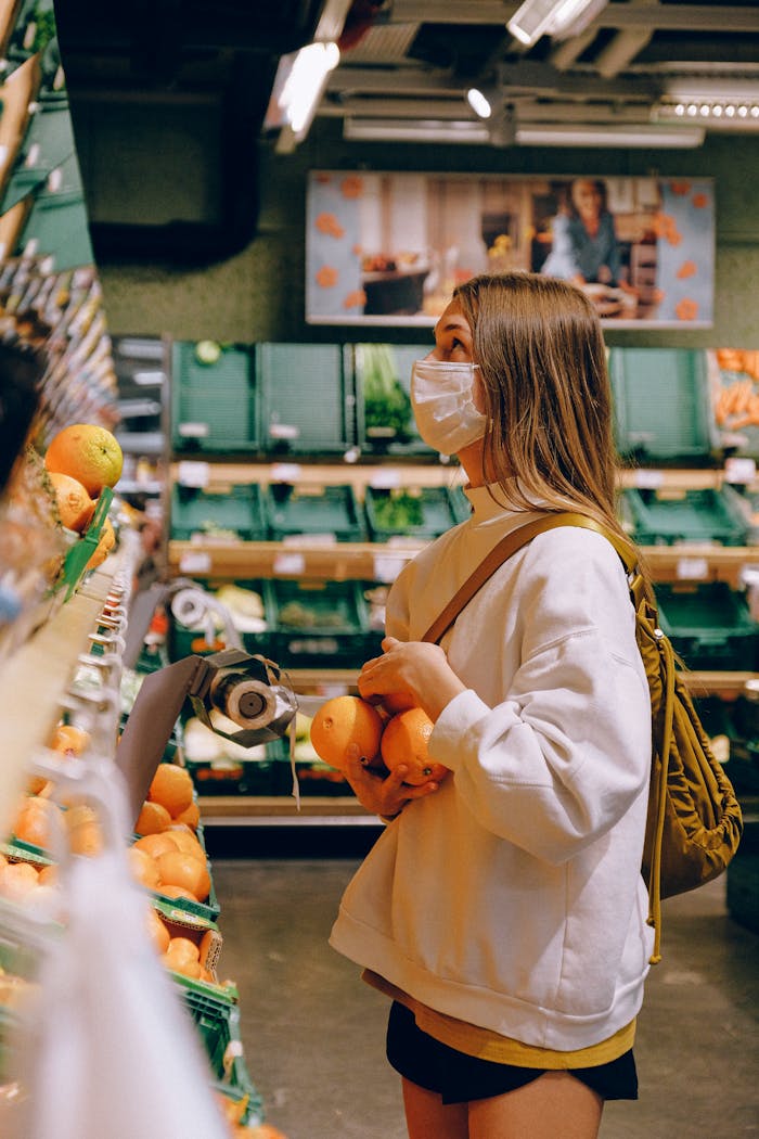 gallery-1 Woman in a mask selects fresh oranges in a supermarket, emphasizing hygiene and protection.