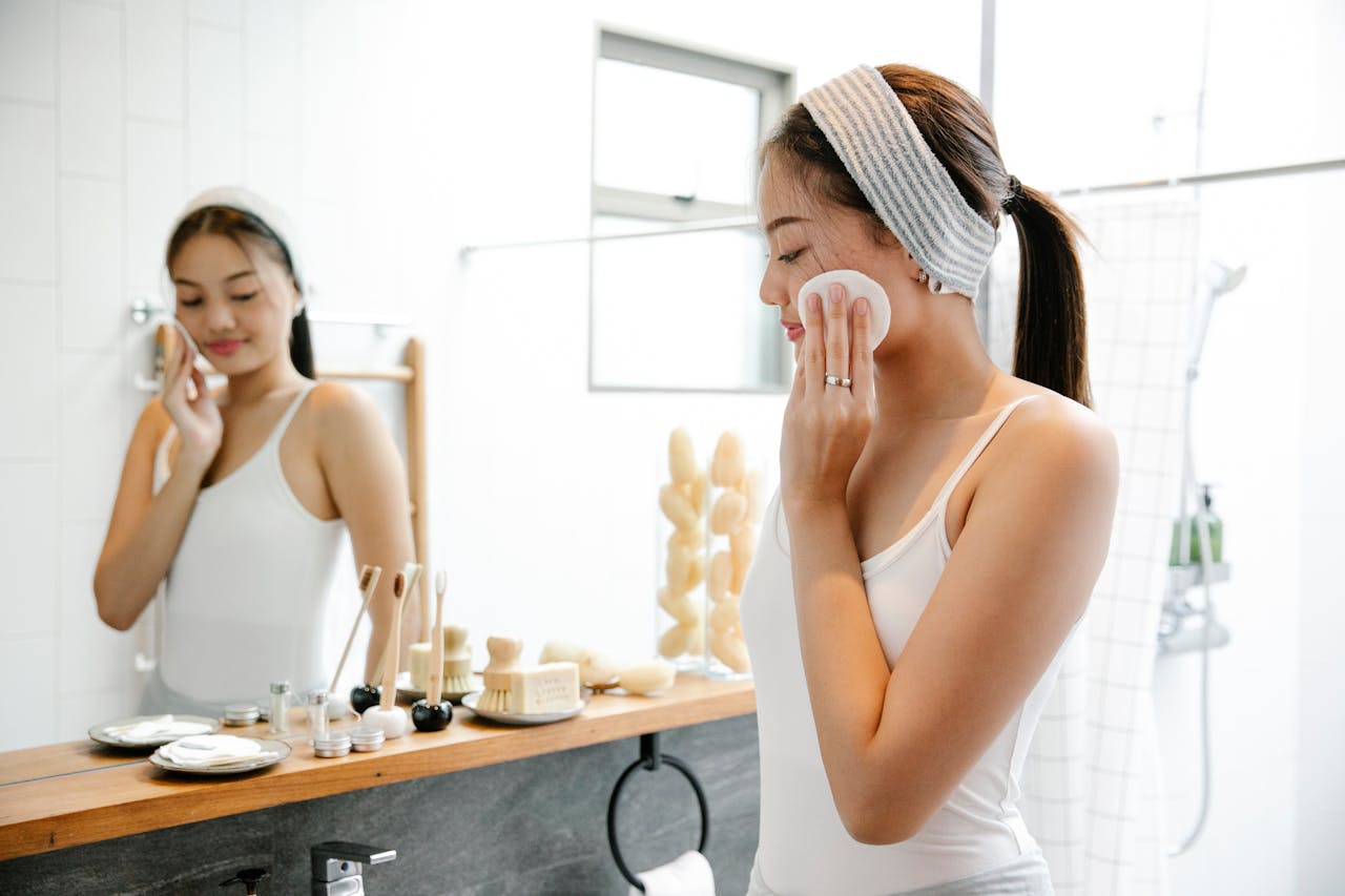 contact-img Asian woman applying skincare with cotton pad in bright bathroom setting.